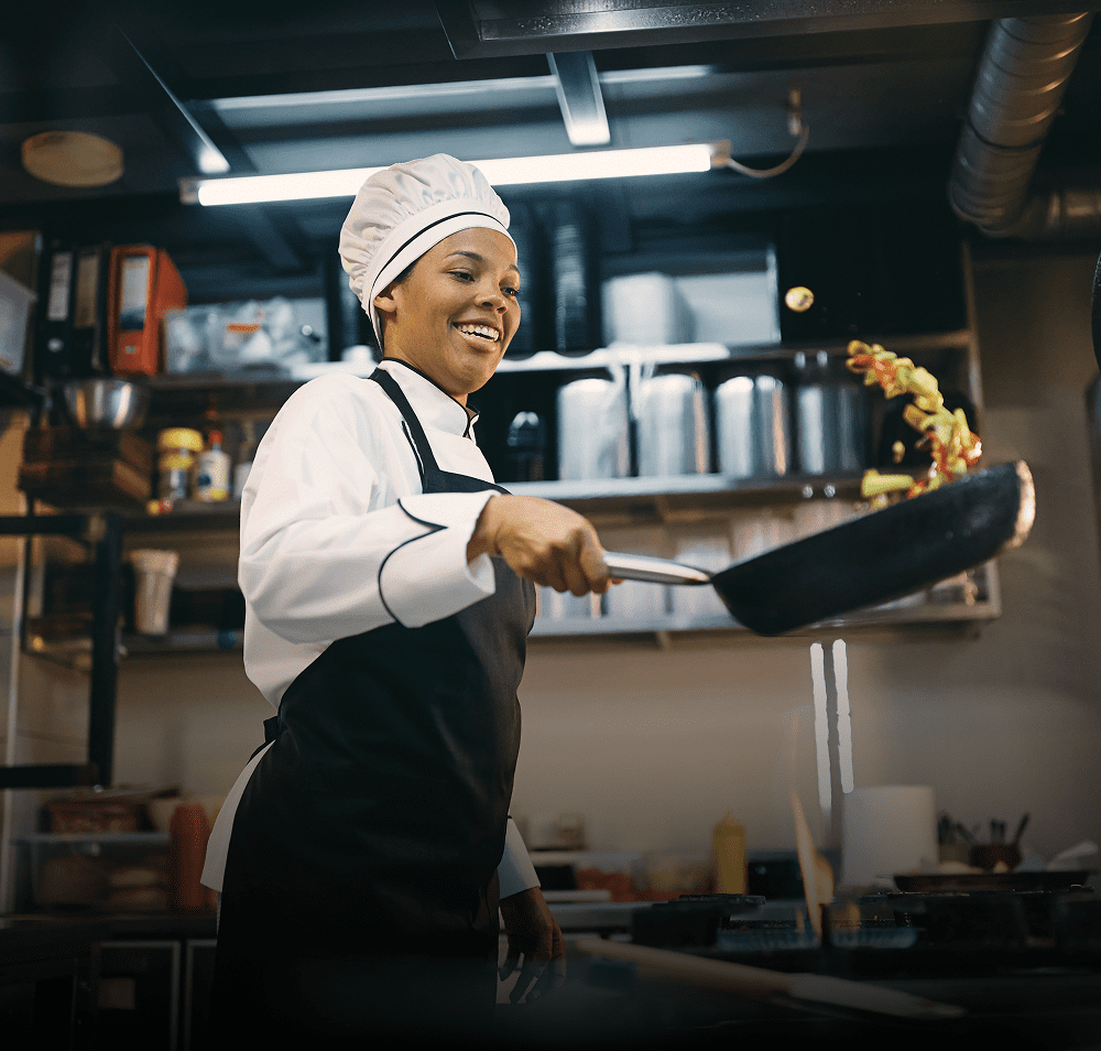 A chef joyfully flips food in a frying pan in a busy kitchen.